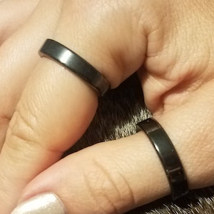 May include: Two black metal rings on a person's fingers. The rings are simple and have a flat, polished surface.