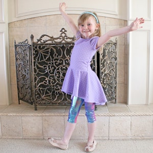 May include: A young girl wearing a lavender dress with white polka dots and iridescent leggings. The dress has a flared skirt and short sleeves. She is wearing a light blue headband and ballet flats. The background includes a fireplace and a decorative screen.