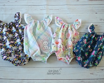 May include: Four baby onesies with different patterns. The onesies have ruffled sleeves with white lace trim. The patterns include a rainbow unicorn design, a plaid pattern with a bunny applique, a floral pattern, and a dark blue floral pattern. The onesies are laid out on a white wooden surface. The text "Sew Sweet, Sew Adorable" is visible at the bottom of the image.
