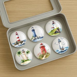 May include: A set of six round magnets featuring watercolor illustrations of lighthouses. The lighthouses are depicted in various colors and settings, including on rocky shores and islands. The magnets are presented in a silver metal tin.