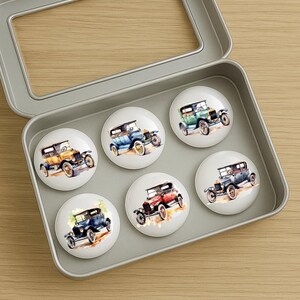May include: A set of six round decorative magnets featuring vintage car illustrations in various colors, including yellow, blue, green, and red. The magnets are displayed in a silver metal tin, perfect for a car enthusiast's collection.