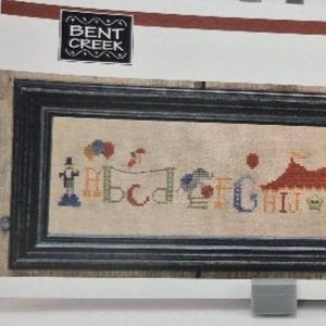 May include: A cross-stitch pattern for a colorful alphabet design with a circus theme. The pattern includes a tent, balloons, and other circus elements. The title of the pattern is "Circus Row".
