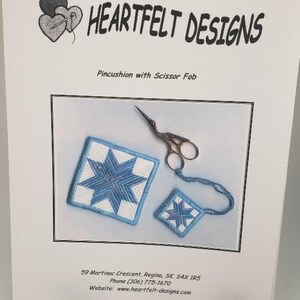 Puede incluir: Un cojín para alfileres azul y blanco con un diseño de estrella y un llavero para tijeras a juego. El cojín para alfileres y el llavero son ambos hechos a mano y presentan un diseño de estrella azul sobre un fondo blanco. El texto "HEARTFELT DESIGNS" está en la parte superior de la imagen. El texto "Pincushion with Scissor Fob" está debajo de la imagen. El texto "59 Martinac Crescent, Regina, SK. S4X 1R5" está en la parte inferior de la imagen. El texto "Phone (306) 775-1670" está debajo de la dirección. El texto "Website: www.heartfelt-designs.com" está debajo del número de teléfono.
