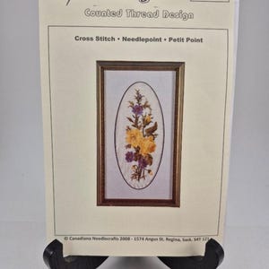 Puede incluir: Un patrón de punto contado para punto de cruz, punto de aguja o punto pequeño. El patrón presenta un óvalo enmarcado con un arreglo floral de flores amarillas y moradas. El patrón se titula "Helen Burgess Counted Thread Design" y está numerado como "506".