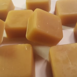 May include: A close-up of ten small, square, golden-brown wax melts. The wax melts are arranged in a random pattern on a white surface.