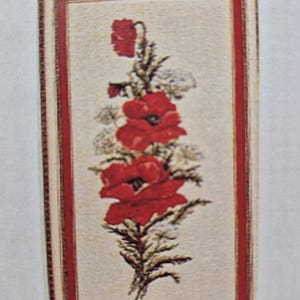 以下が含まれることがあります： 赤いポピーの花束を緑色の茎と白い花で刺繍した、額装されたクロスステッチ刺繍。刺繍はクリーム色の背景に施されています。
