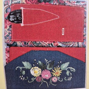 May include: A handmade wallet with a red corduroy exterior and a floral paisley interior. The wallet features a red pocket with a black item inside, and a blue denim pocket with embroidered flowers.