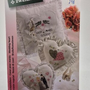 May include: Three white heart-shaped pillows with lace trim, each embroidered with a wedding theme. The pillows feature the names "Lilly & Marc" and "Marc & Lilly" with wedding dates. The pillows are surrounded by white doves and a bouquet of pink and white roses.