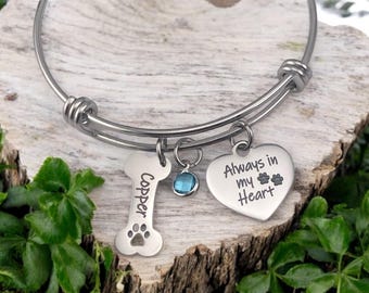 May include: A silver expandable bracelet with three charms: a bone charm engraved with the name "Copper", a heart charm engraved with "Always in my Heart" and paw prints, and a small blue crystal charm.