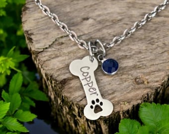 May include: A silver dog bone charm necklace with a blue gemstone charm. The bone charm is engraved with the name "Copper".