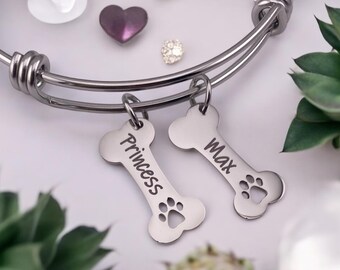 May include: A silver-toned expandable bracelet with two bone-shaped charms. The charms are engraved with the names "Princess" and "Max" and feature paw print cutouts. Decorative elements include a purple heart and a small, sparkly gem.