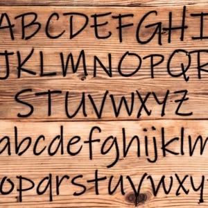 May include: A close-up of a wooden surface with the alphabet written in black marker. The letters are written in a casual, handwritten style. The letters are arranged in three rows, with the uppercase letters on the top row, the lowercase letters on the second row, and the lowercase letters on the third row.