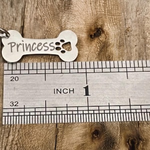 May include: A silver bone-shaped dog tag with the name "Princess" engraved on it. The tag is 1 inch long and has a paw print cutout.