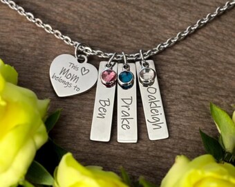 May include: A silver necklace with a heart-shaped pendant engraved with "This Mom belongs to" and three rectangular charms with names and gemstones. The necklace is displayed with yellow roses.