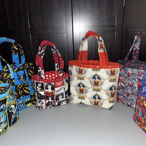 May include: Seven colorful tote bags with superhero themes. The bags are made of fabric and have different designs featuring Batman, Wonder Woman, and Spiderman. The bags have black, blue, red, and yellow fabric.