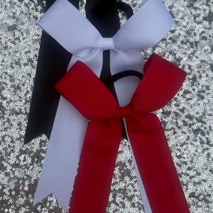 May include: Three hair bows made of grosgrain ribbon, each attached to a black elastic hair tie. The bows are stacked vertically in black, white, and red. Each bow features two long, pointed tails extending downward from the central knot.