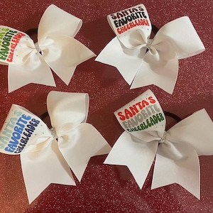 Pode incluir: Quatro laços de torcida brancos com um design "Santa's Favorite Cheerleader" em vermelho, verde, azul e cinza. Os laços são amarrados com uma fita branca e têm um strass no centro.