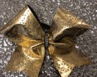 May include: A gold cheer bow with rhinestones. The bow is on a silver sequin background.