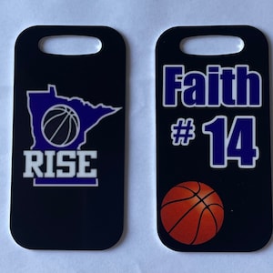 May include: Two black luggage tags with a basketball theme. The first tag has a basketball inside the outline of the state of Minnesota with the word "RISE" below. The second tag has the name "Faith" and the number "14" in blue with a basketball in the lower right corner.