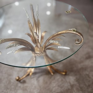May include: A small, round glass-topped table with a gold-colored metal base. The base features an ornate, stylized floral design with curved elements and decorative accents. The table is set on a neutral-toned carpet.