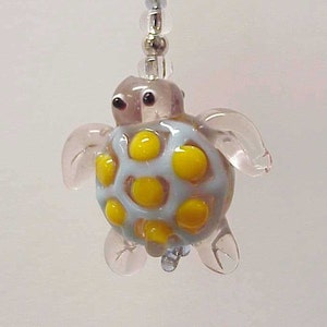 May include: A small, glass turtle pendant with a light blue shell featuring yellow spots. The turtle has clear flippers and head, with black eyes. It hangs from a beaded strand, creating a whimsical charm.