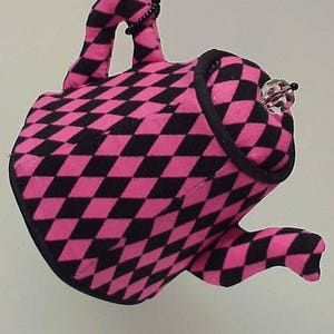 May include: A pink and black checkered teapot with a crystal-like ornament hanging from the spout. The teapot is made of fabric and has a whimsical, handmade look.