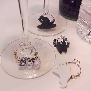 May include: Four Halloween-themed wine glass charms. One charm features a black witch's hat, another a black bat, another the word "BOO" in white with black accents, and the last a white ghost.