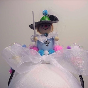 May include: A white tulle bow with a small doll wearing a blue dress and a green hat. The doll is holding a white wand. The bow is decorated with pink, blue, and white pom-poms.