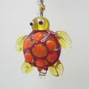 May include: A yellow glass turtle charm with a red and orange shell. The turtle has small black eyes and is hanging from a string of beads.