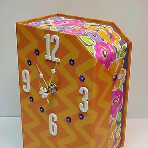 May include: A decorative clock with an orange chevron pattern on the front and sides. The clock face has white numbers and hands, with small blue flower accents. The top and side edges feature a floral design with pink, orange, and purple flowers.