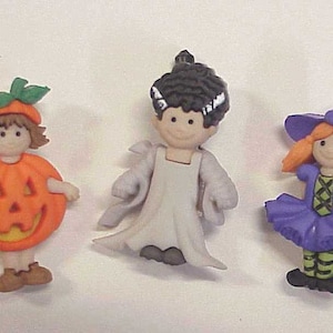 May include: Three Halloween-themed figurines: a pumpkin, a bride of Frankenstein, and a witch. The pumpkin figure is orange with a green stem, the bride is in a white dress, and the witch wears a purple hat and dress.