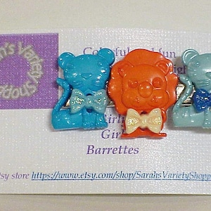 May include: Three colorful plastic animal barrettes. A blue cat, an orange lion, and a teal cat. Each barrette has a bow tie. The barrettes are on a white background with the text "Colorful Fun Girl Barrettes" and "Etsy store https://www.etsy.com/shop/SarahsVarietyShoppe".