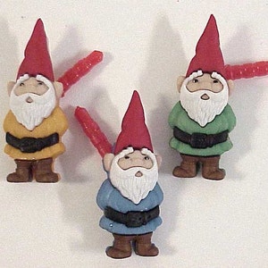 May include: Three small gnome figurines with red hats and white beards. The gnomes are wearing yellow, green, and blue outfits. Each gnome has a red and white striped stick attached to its back.