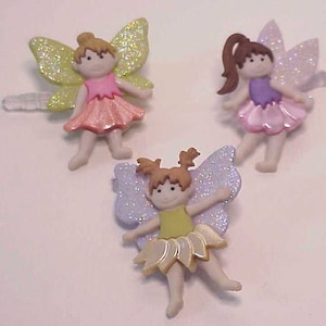 May include: Three fairy hair clips with glitter wings. The fairies are wearing pink, purple, and yellow dresses.