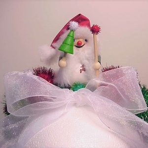 May include: A white snowman figurine with a red hat and a green Christmas tree in its hand. The snowman is sitting on a white fluffy base with a white sheer bow and green and red tinsel.