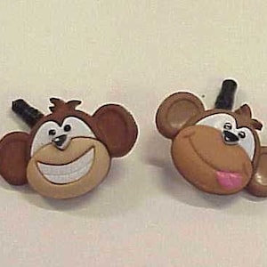 May include: Four cartoon monkey face charms with brown, tan, and white details. Each charm has a different facial expression, including a smile, a tongue sticking out, and a neutral expression. The charms have black accents.
