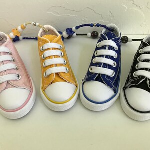 May include: Four miniature canvas sneakers with white laces. The sneakers are yellow, pink, blue, and black. The sneakers are connected by a beaded chain.