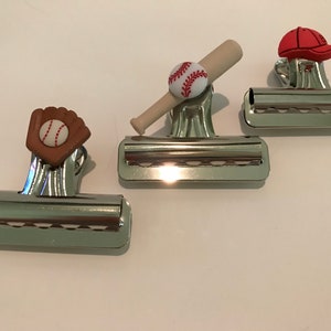 May include: Three silver binder clips with baseball-themed embellishments. One clip has a baseball and bat, one has a baseball glove, and one has a red baseball cap.