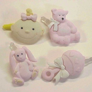 May include: Four pink and white baby themed plastic charms with a yellow, pink, and white bow, a pink bear, a pink bunny, and a pink rattle.