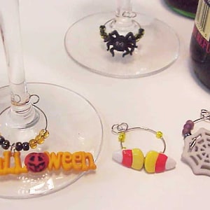 May include: Four Halloween themed wine glass charms. One charm features a black spider with yellow beads, another has a white spider web, another has a candy corn design, and the last charm has a pumpkin and the word "Halloween" in orange letters.