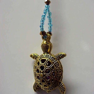 May include: A gold-colored turtle pendant with a detailed shell design and colorful gemstones. The pendant is attached to a blue beaded chain.