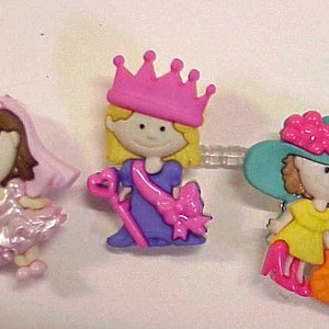 May include: Three plastic hair clips featuring cartoon girls. The first clip has a pink dress and a pink crown. The second clip has a purple dress and a pink crown. The third clip has a yellow dress and a blue hat.