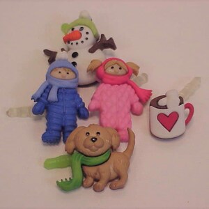 May include: A collection of winter-themed buttons. Includes a snowman, two figures in snowsuits, a dog with a green scarf, and a mug with a heart. The buttons are in various colors, including blue, pink, brown, and white.