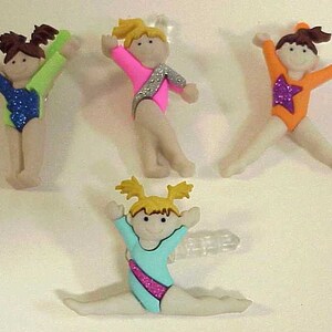 May include: Four colorful plastic figurines of gymnasts in various poses. Each gymnast is wearing a sparkly leotard and has blonde hair with pigtails.
