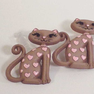 May include: Two brown ceramic cat buttons with pink hearts. The cats have black eyes and whiskers.