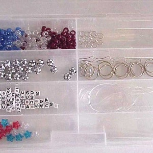 May include: A clear plastic storage box with multiple compartments filled with various beads, charms, and jump rings. The beads are in red, white, and blue colors. The charms are in the shape of soccer balls and letters. The jump rings are silver.