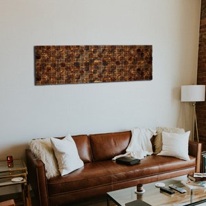 Op de afbeelding: Een bruine leren bank met witte kussens en een deken staat in een woonkamer met een houten wand en een groot raam. Een glazen salontafel staat voor de bank met een boek en een afstandsbediening erop. Een koeienhuidkleed ligt op de vloer.