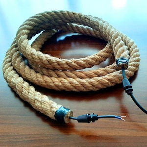 Puede incluir: Un tramo de cuerda de cáñamo natural enrollado con cable eléctrico negro y conectores en cada extremo. La cuerda es de color marrón claro y el cable es negro.