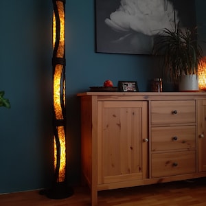 K&ouml;nnte beinhalten: Eine hohe, schwarze Stehlampe mit einem warmen, gelben Licht, das aus dem Inneren strahlt. Die Lampe ist aus gewebtem Material gefertigt und hat eine zylindrische Form. Die Lampe steht auf einem Holzboden vor einem Holzschrank mit Schubladen.