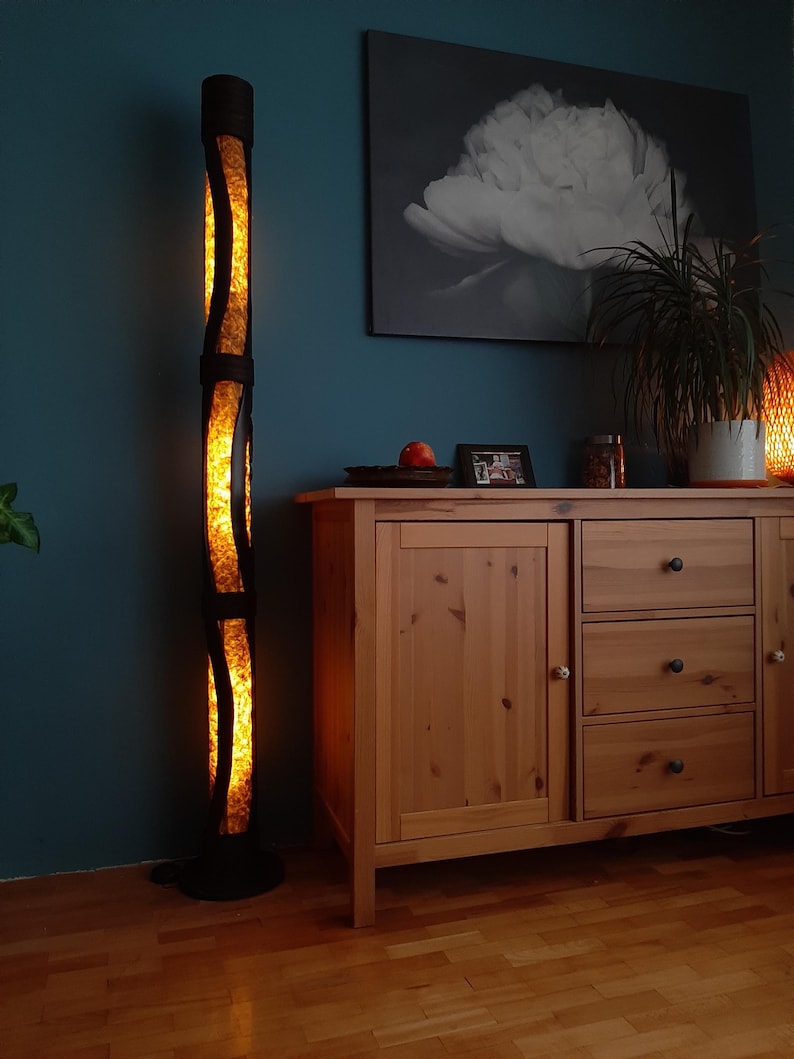 K&ouml;nnte beinhalten: Eine hohe, schwarze Stehlampe mit einem warmen, gelben Licht, das aus dem Inneren strahlt. Die Lampe ist aus gewebtem Material gefertigt und hat eine zylindrische Form. Die Lampe steht auf einem Holzboden vor einem Holzschrank mit Schubladen.